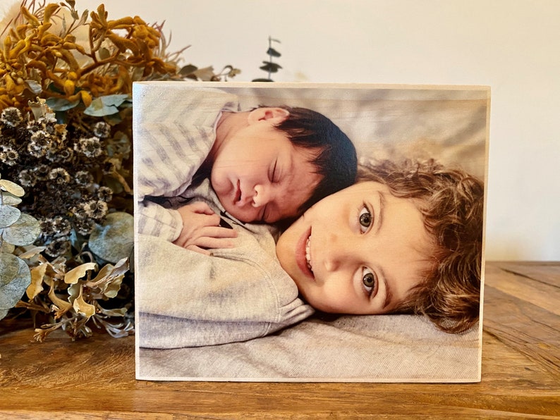 May include: A close-up photo of two siblings. An older child with curly brown hair is lying down with a younger baby sibling sleeping on their chest. The baby is wearing a striped shirt and the older child is wearing a grey shirt.