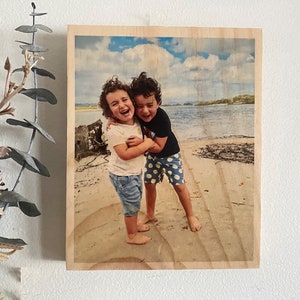 May include: Two children, a girl and a boy, are laughing and hugging on a sandy beach. The girl is wearing a white shirt, blue jeans, and has curly brown hair. The boy is wearing a black shirt, blue and white polka dot shorts, and has brown hair.