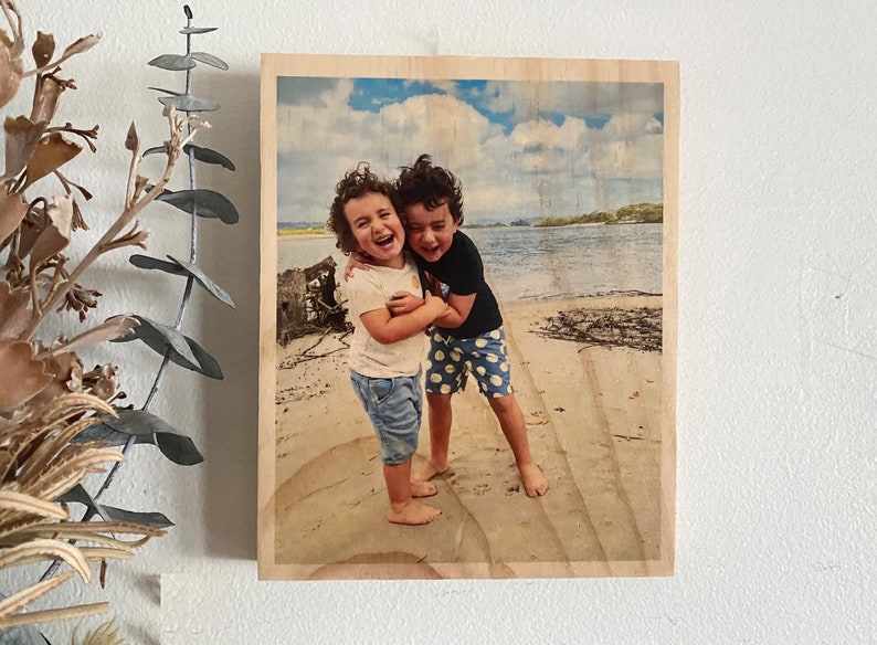 May include: Two children, a girl and a boy, are laughing and hugging on a sandy beach. The girl is wearing a white shirt, blue jeans, and has curly brown hair. The boy is wearing a black shirt, blue and white polka dot shorts, and has brown hair.
