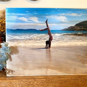 May include: A young person doing a handstand on a sandy beach with blue water and a cloudy sky in the background.