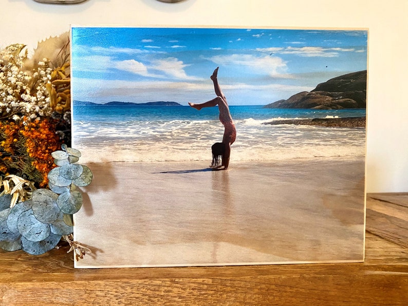 May include: A young person doing a handstand on a sandy beach with blue water and a cloudy sky in the background.