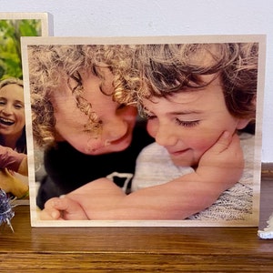 May include: A wooden photo block with a printed image of two children hugging. The children have curly hair and are smiling. The photo block is a light brown color.