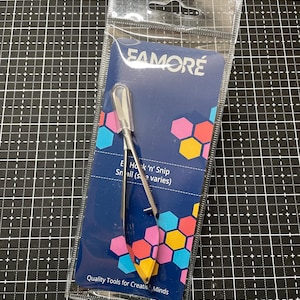 May include: A silver metal Famore E Hook 'n' Snip tool with a clear plastic protective cap, packaged in a clear plastic sleeve with a blue backing card featuring colorful hexagon patterns and the text Quality Tools for Creative Minds.
