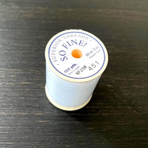 May include: A spool of light blue polyester thread with a white label reading Superior Threads So Fine! 50 wt. 3-ply, 550 yds., Made in Japan, WF25R, and 451. The thread is wound around a cylindrical plastic core with a central orange opening.
