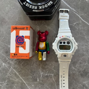 May include: A white G-Shock digital watch with a black metal tin, a Series 15 Bearbrick figure, and a colorful Bearbrick figure. The watch has a white band with the text "APE SHALL NEVER KILL APE". The tin says "SHOCK RESIST".