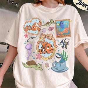 Könnte beinhalten: Ein elfenbeinfarbenes T-Shirt mit einem farbenfrohen Grafikdruck verschiedener Cartoon-Meeresbewohner, darunter Clownfische, eine Meeresschildkröte, ein Rochen, eine Qualle und ein Hai in einer Teetasse, mit dem Text Just Keep Swimming.