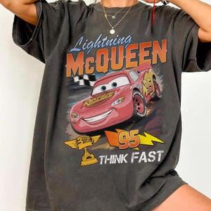 May include: Dark gray t-shirt featuring Lightning McQueen from the Cars movie. The graphic includes McQueen, the number 95, and the words "THINK FAST". The shirt has a vintage, distressed look.