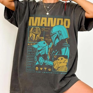 May include: A charcoal gray short-sleeve t-shirt featuring a graphic print with the text MANDO AND GROGU. The design includes illustrations of the characters in yellow and teal, with the phrase WHEREVER I GO, HE GOES and THE LEGEND CONTINUES.