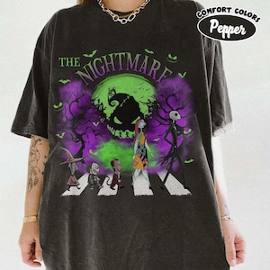 Pu&ograve; includere: T-shirt grigio scuro con grafica di "Nightmare Before Christmas". Il design mostra personaggi che attraversano una strada con una luna verde e accenti viola. Il testo "THE NIGHTMARE" &egrave; arcuato nella parte superiore.