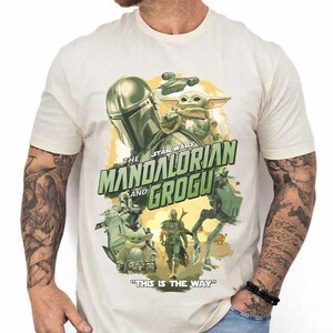May include: Cream-colored t-shirt featuring a graphic from the Star Wars series, "The Mandalorian and Grogu." The design includes images of the Mandalorian, Grogu, and other characters, with the phrase "This is the Way."