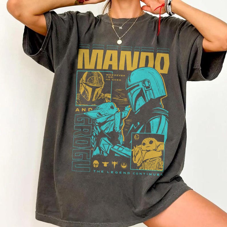 May include: A charcoal gray short-sleeve t-shirt featuring a graphic print with the text MANDO AND GROGU. The design includes illustrations of the characters in yellow and teal, with the phrase WHEREVER I GO, HE GOES and THE LEGEND CONTINUES.