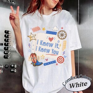 May include: A white t-shirt featuring a central graphic of a lined notepad page with the blue text I Knew It, I Knew You. The design includes a red heart, a brown cowboy boot, a sheriff star, a wagon wheel, and various small colorful shapes.