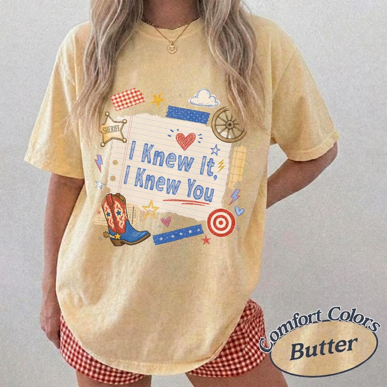 May include: A pale yellow short-sleeve t-shirt featuring a graphic print of a lined notebook page with the text I Knew It, I Knew You. The design includes a red heart, a blue cowboy boot, a sheriff star, a wagon wheel, and small stars.