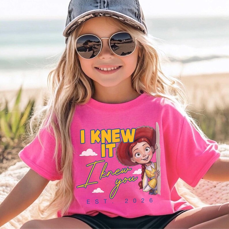 May include: A bright pink short-sleeved t-shirt featuring a graphic of the character Jessie from Toy Story. The text reads I KNEW IT I knew you EST. 2026 in yellow and blue lettering, accompanied by small white cloud illustrations.