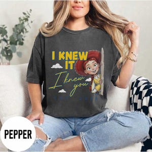 May include: A charcoal gray short-sleeve t-shirt featuring a graphic of a cartoon character with red hair. The text reads I KNEW IT in yellow and I knew you in cursive yellow script, with small white clouds and the text EST 2036 in blue.