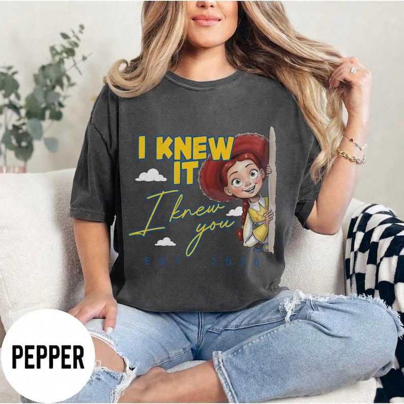 May include: A charcoal gray short-sleeve t-shirt featuring a graphic of a cartoon character with red hair. The text reads I KNEW IT in yellow and I knew you in cursive yellow script, with small white clouds and the text EST 2036 in blue.
