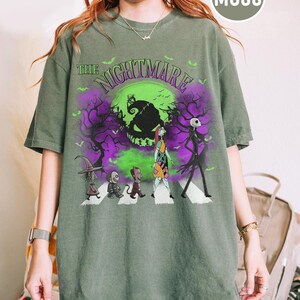 Pu&ograve; includere: T-shirt verde muschio con grafica di "Nightmare Before Christmas". Il design include Jack Skellington, Sally e altri personaggi che attraversano le strisce pedonali davanti a una luna verde e un cielo viola. Il testo "The Nightmare" &egrave; sopra.