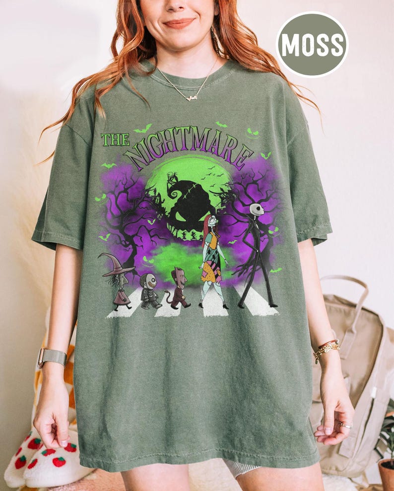 Pu&ograve; includere: T-shirt verde muschio con grafica di "Nightmare Before Christmas". Il design include Jack Skellington, Sally e altri personaggi che attraversano le strisce pedonali davanti a una luna verde e un cielo viola. Il testo "The Nightmare" &egrave; sopra.