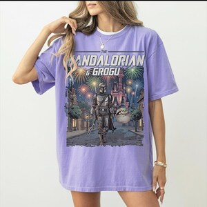 Op de afbeelding: Een lichtpaars t-shirt met korte mouwen en een grafische print van The Mandalorian en Grogu die over een straat lopen met een kasteel op de achtergrond onder vuurwerk. De tekst THE MANDALORIAN & GROGU staat boven de illustratie.