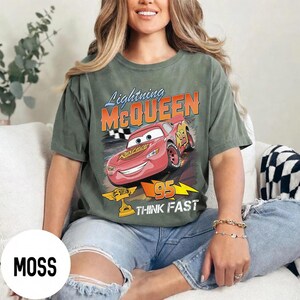 May include: Moss green t-shirt featuring a graphic of Lightning McQueen from the Cars movie. The design includes the text "Lightning McQueen", "95", and "Think Fast". The shirt is a relaxed fit and has short sleeves.