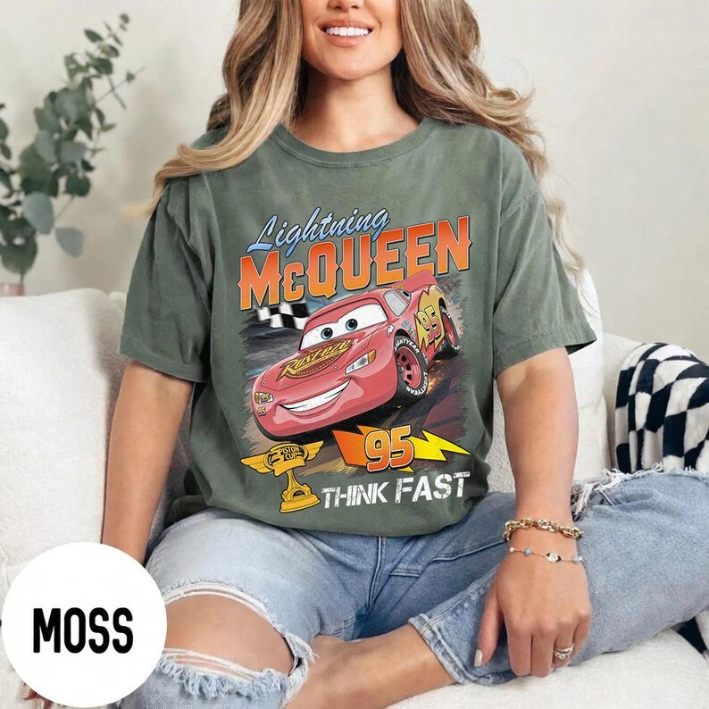 May include: Moss green t-shirt featuring a graphic of Lightning McQueen from the Cars movie. The design includes the text "Lightning McQueen", "95", and "Think Fast". The shirt is a relaxed fit and has short sleeves.