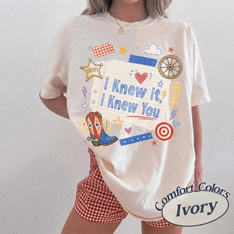 May include: An ivory t-shirt featuring a central graphic of a lined notebook page with the text "I Knew It, I Knew You" in blue. The design includes a cowboy boot, a sheriff star, a wagon wheel, a target, and various colorful doodles.