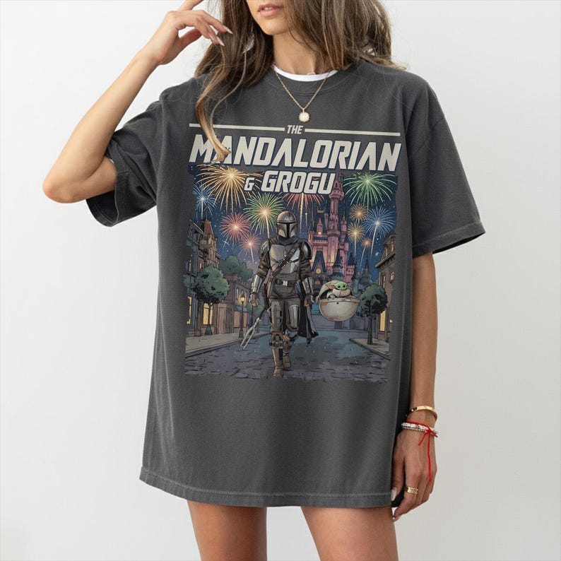 Op de afbeelding: Een antracietgrijs t-shirt met een grafische print van The Mandalorian en Grogu die over een straat lopen met vuurwerk aan de nachtelijke hemel boven een kasteel. De tekst THE MANDALORIAN & GROGU staat in het wit bovenaan de afbeelding.