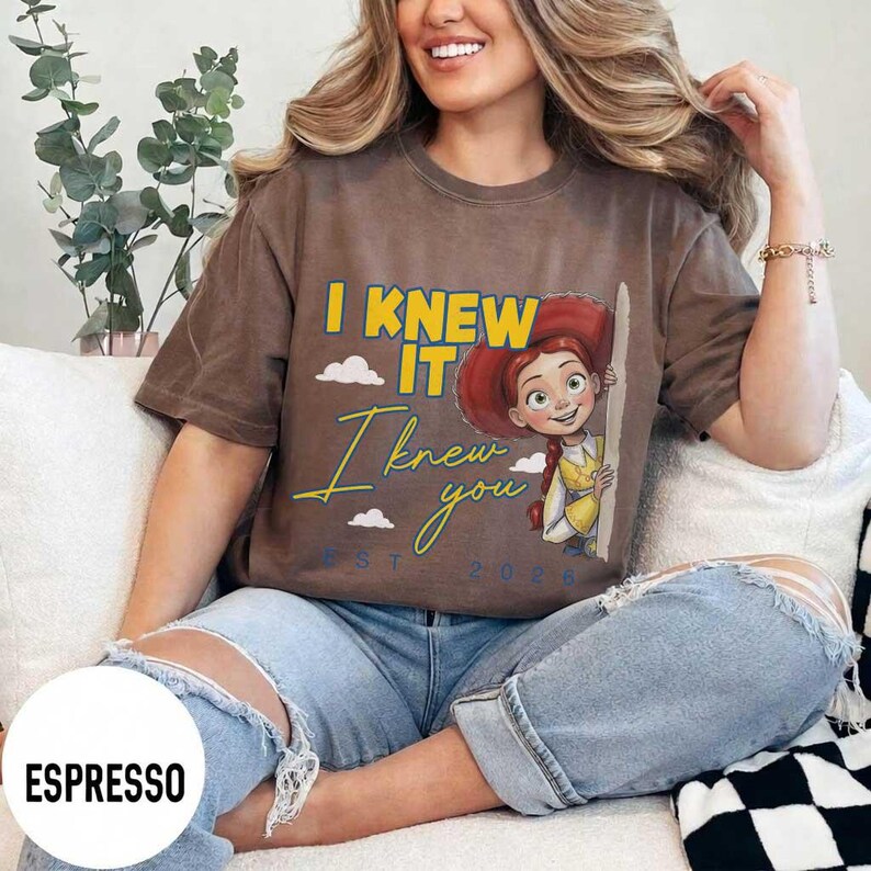 May include: A brown short-sleeved t-shirt featuring a graphic of a cartoon cowgirl character with red hair. The text reads I KNEW IT in yellow, I knew you in cursive yellow, and EST. 2026 in blue. Small white clouds are printed around the character.