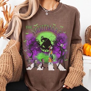 Pu&ograve; includere: T-shirt marrone con grafica di "Nightmare Before Christmas". Il design mostra personaggi che attraversano le strisce pedonali davanti a una luna verde e nebbia viola. Il testo "The Nightmare" &egrave; arcuato sopra.