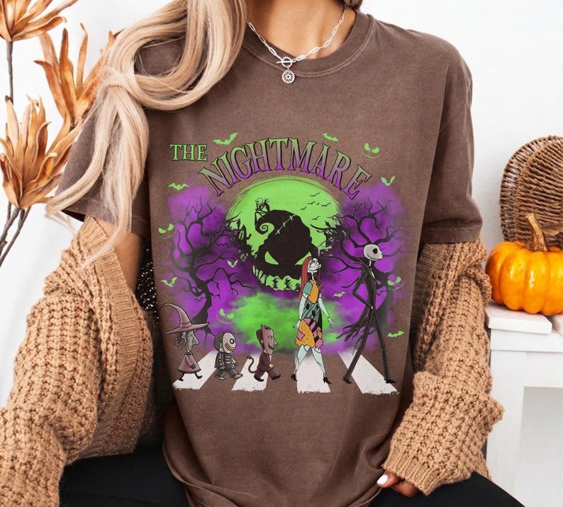 Pu&ograve; includere: T-shirt marrone con grafica di "Nightmare Before Christmas". Il design mostra personaggi che attraversano le strisce pedonali davanti a una luna verde e nebbia viola. Il testo "The Nightmare" &egrave; arcuato sopra.
