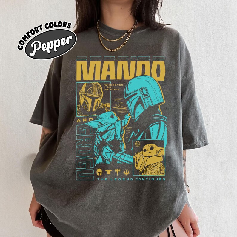 May include: A charcoal gray short-sleeve t-shirt featuring a graphic print of Mando and Grogu. The design includes yellow and teal text reading MANDO AND GROGU, WHEREVER I GO, HE GOES, and THE LEGEND CONTINUES, along with small character icons.