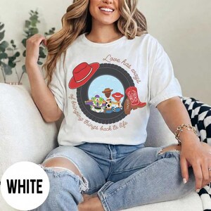 Pu&ograve; includere: Una t-shirt bianca con una grafica raffigurante uno pneumatico nero che incornicia personaggi dei cartoni animati, un cappello da cowboy rosso e uno stivale da cowboy rosso. Il testo Love has ways of bringing things back to life circonda l'immagine.