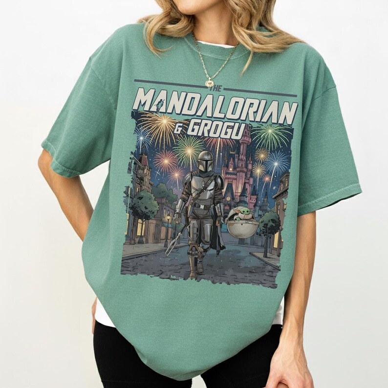 Op de afbeelding: Een saliegroen t-shirt met een grafische print van The Mandalorian en Grogu die 's nachts door een straat lopen. Het ontwerp bevat een kasteel op de achtergrond, kleurrijk vuurwerk in de lucht en de tekst THE MANDALORIAN & GROGU.