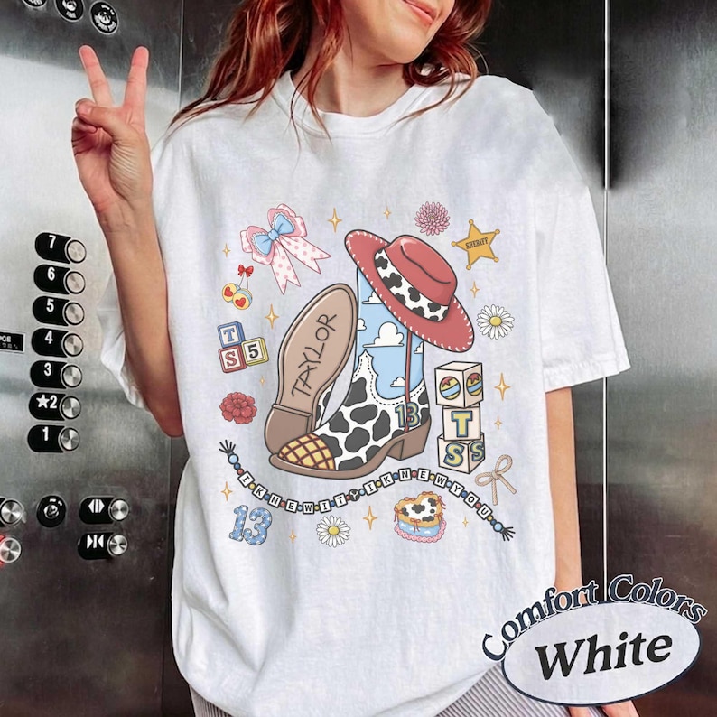 Op de afbeelding: Een wit t-shirt met een grafische print van een cowboylaars met koeienvlekken, een rode cowboyhoed, alfabetblokken met de letters T, S en 5, een roze strik, een ster en een kralenketting. Het woord TAYLOR staat op de zool.