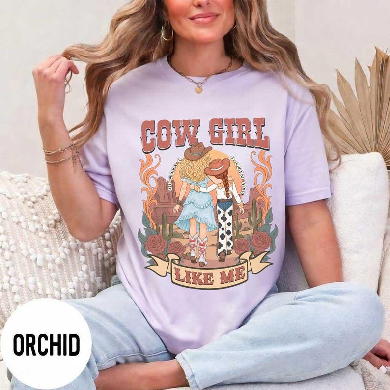 May include: A light purple t-shirt featuring a graphic print of two figures in cowboy hats standing in a desert landscape with cacti and roses. The text "COW GIRL" appears above the figures, and "LIKE ME" is printed on a banner below them.
