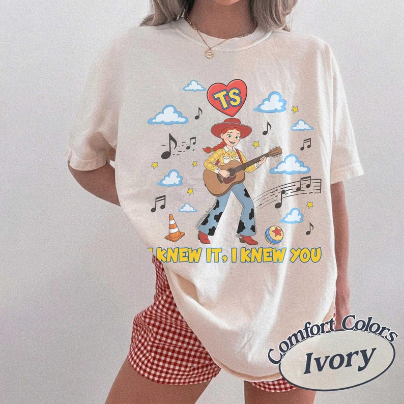 May include: An ivory t-shirt featuring a graphic of a cowgirl playing an acoustic guitar, surrounded by blue clouds, musical notes, a traffic cone, a ball, and a red heart with the letters TS. The text I KNEW IT, I KNEW YOU is printed in yellow.