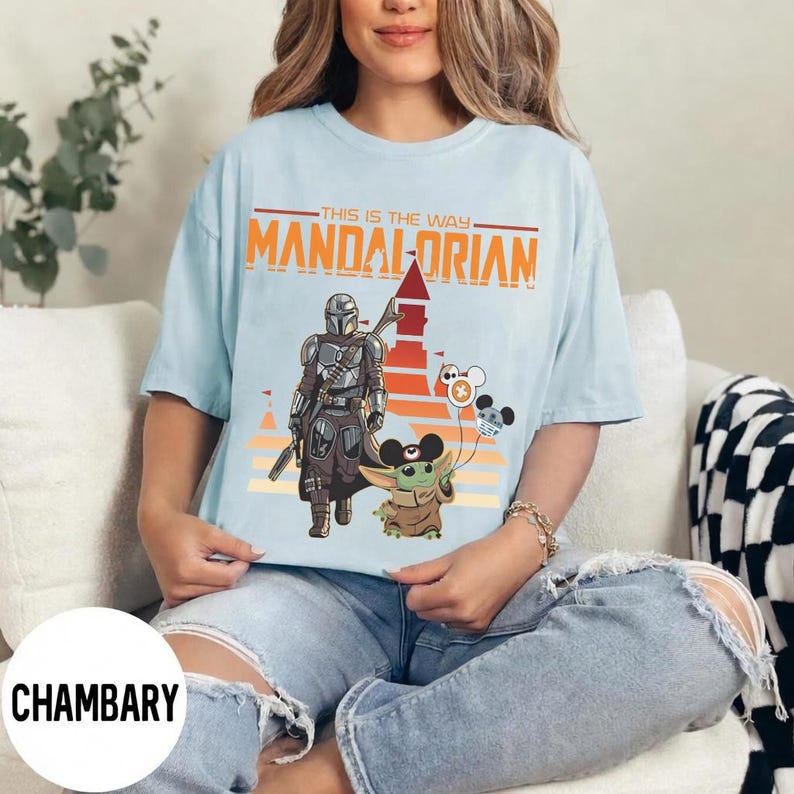 May include: Light blue t-shirt featuring a graphic of The Mandalorian and Grogu, with the text "THIS IS THE WAY MANDALORIAN". The design includes a sunset-colored background and Mickey Mouse-themed balloons. The shirt is a relaxed fit.