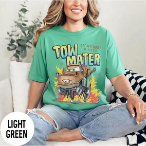 May include: A light green t-shirt featuring a graphic of the character Tow Mater from the movie Cars. The text reads Tow Mater and Off Without A Hitch. The design includes a brown tow truck with large wheels surrounded by yellow and orange flames.