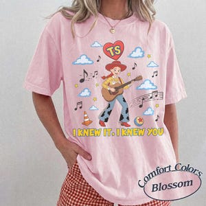 May include: A light pink t-shirt featuring a graphic of a cowgirl playing an acoustic guitar, surrounded by blue clouds, musical notes, a traffic cone, a ball, and a red heart with the letters TS. The text I KNEW IT, I KNEW YOU is printed below.
