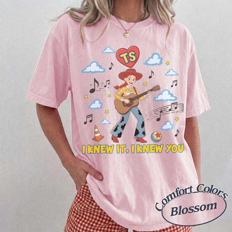 May include: A light pink t-shirt featuring a graphic of a cowgirl playing an acoustic guitar, surrounded by blue clouds, musical notes, a traffic cone, a ball, and a red heart with the letters TS. The text I KNEW IT, I KNEW YOU is printed below.