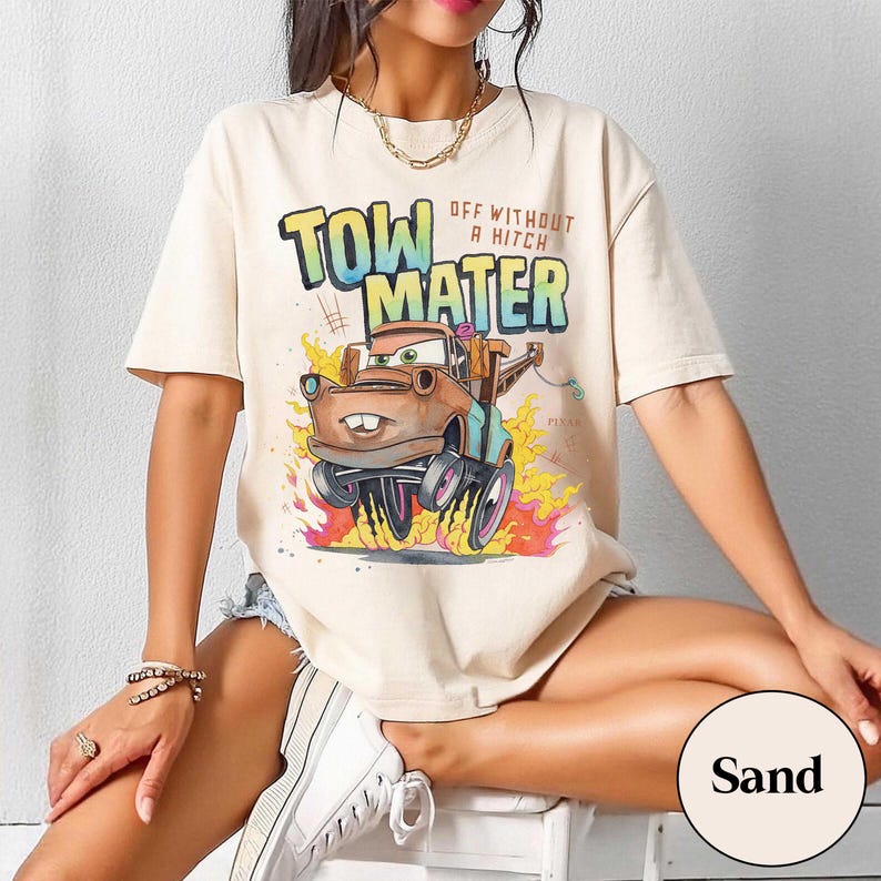 May include: A sand-colored short-sleeve t-shirt featuring a graphic of the character Tow Mater from the movie Cars. The text reads Tow Mater and Off Without A Hitch. The design includes flames and a colorful gradient effect on the lettering.