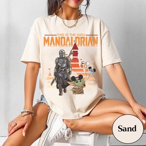 May include: Sand-colored t-shirt featuring a graphic of The Mandalorian and Grogu, with the text "THIS IS THE WAY" and "MANDALORIAN" in orange. Grogu holds balloons with Mickey Mouse ears. The design is set against a sunset backdrop.