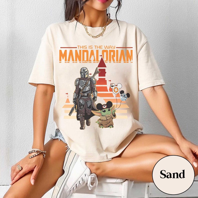 May include: Sand-colored t-shirt featuring a graphic of The Mandalorian and Grogu, with the text "THIS IS THE WAY" and "MANDALORIAN" in orange. Grogu holds balloons with Mickey Mouse ears. The design is set against a sunset backdrop.