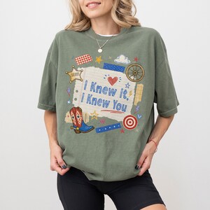 May include: An olive green short-sleeve t-shirt featuring a central graphic on a lined paper background with the text I Knew It, I Knew You. The design includes a cowboy boot, a sheriff star, a wagon wheel, a target, and various small stars.