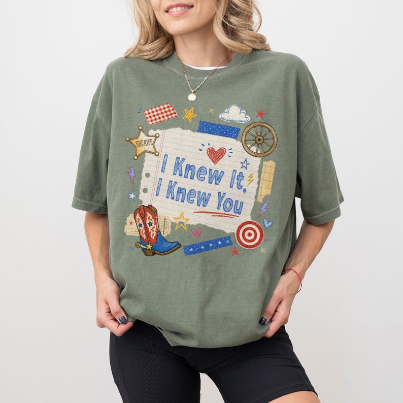May include: An olive green short-sleeve t-shirt featuring a central graphic on a lined paper background with the text I Knew It, I Knew You. The design includes a cowboy boot, a sheriff star, a wagon wheel, a target, and various small stars.