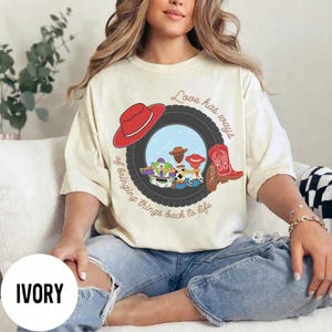 Pu&ograve; includere: Una t-shirt color avorio con una grafica di uno pneumatico nero che incornicia personaggi dei cartoni animati, un cappello da cowboy rosso e uno stivale da cowboy rosso. Il testo recita Love has ways of bringing things back to life, con Taylor sullo stivale.
