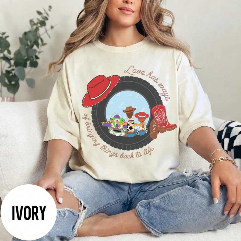 Pu&ograve; includere: Una t-shirt color avorio con una grafica di uno pneumatico nero che incornicia personaggi dei cartoni animati, un cappello da cowboy rosso e uno stivale da cowboy rosso. Il testo recita Love has ways of bringing things back to life, con Taylor sullo stivale.