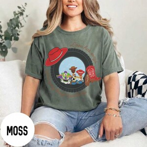 Pu&ograve; includere: Una t-shirt verde muschio con la grafica di uno pneumatico nero che incornicia una scena con i personaggi dei cartoni animati Buzz Lightyear, Woody e Jessie. Il design include un cappello da cowboy rosso, uno stivale rosso e il testo Love has ways e Taylor.