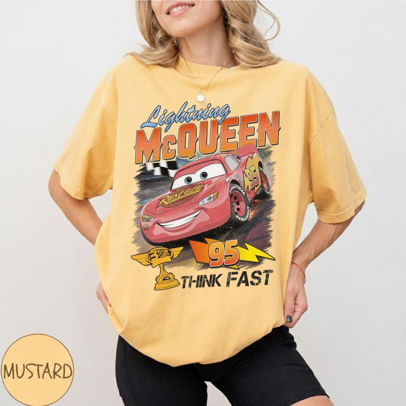 May include: Mustard-colored t-shirt featuring a graphic of Lightning McQueen from the movie Cars. The design includes the text "Lightning McQueen", "95", and "Think Fast". The shirt has short sleeves and a crew neck.
