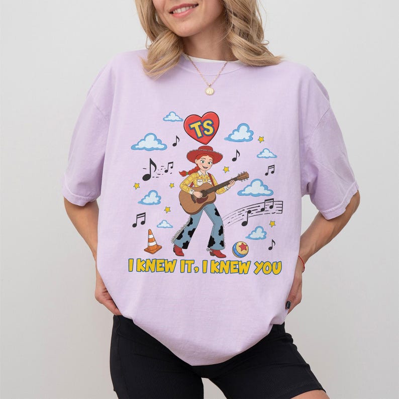 May include: A light purple t-shirt featuring a graphic of a cowgirl playing an acoustic guitar. The design includes a red heart with the letters TS, blue clouds, musical notes, stars, a traffic cone, a ball, and the text I KNEW IT, I KNEW YOU.